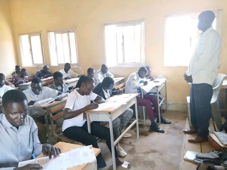 Learners sit at desks while an adult stands at the front of a classroom