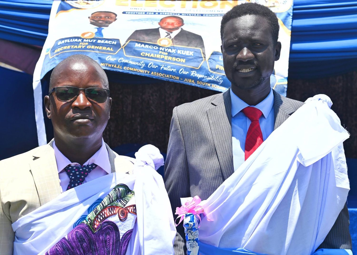 Community event in Juba with a banner naming Marco Nyak Kuek for chairperson; two attendees pose in ceremonial cloth