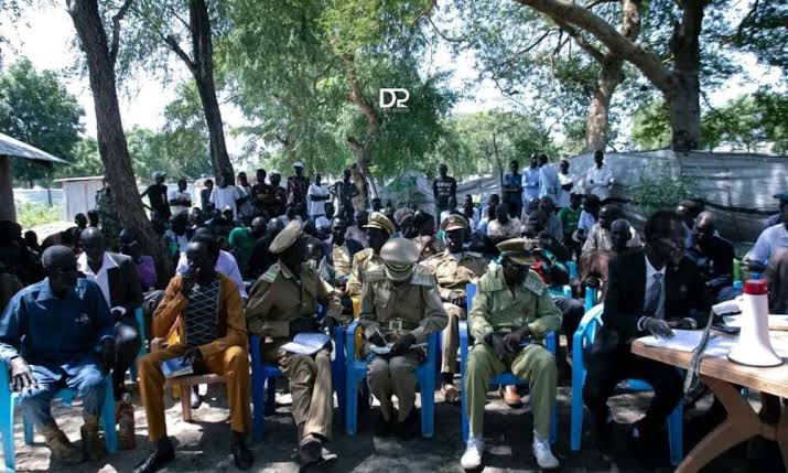 People gathered outdoors beneath trees, with several uniformed participants seated at the front